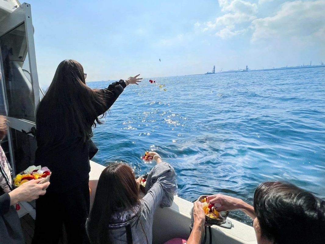人々がボートから青い海に花びらを撒いている様子。晴れた空の下、水面には太陽の光が反射し、遠くには工業地帯のような風景が見えます。故人を偲ぶ海洋散骨のセレモニーのようです。