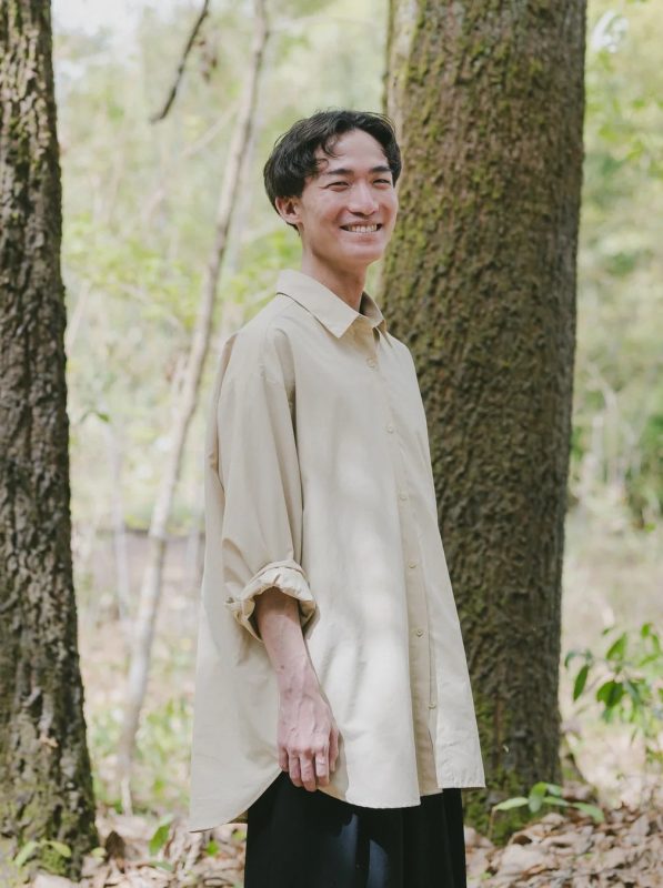 森の中でベージュのシャツを着た男性が優しい笑顔を見せているポートレート写真です。自然光が差し込む穏やかな雰囲気です。