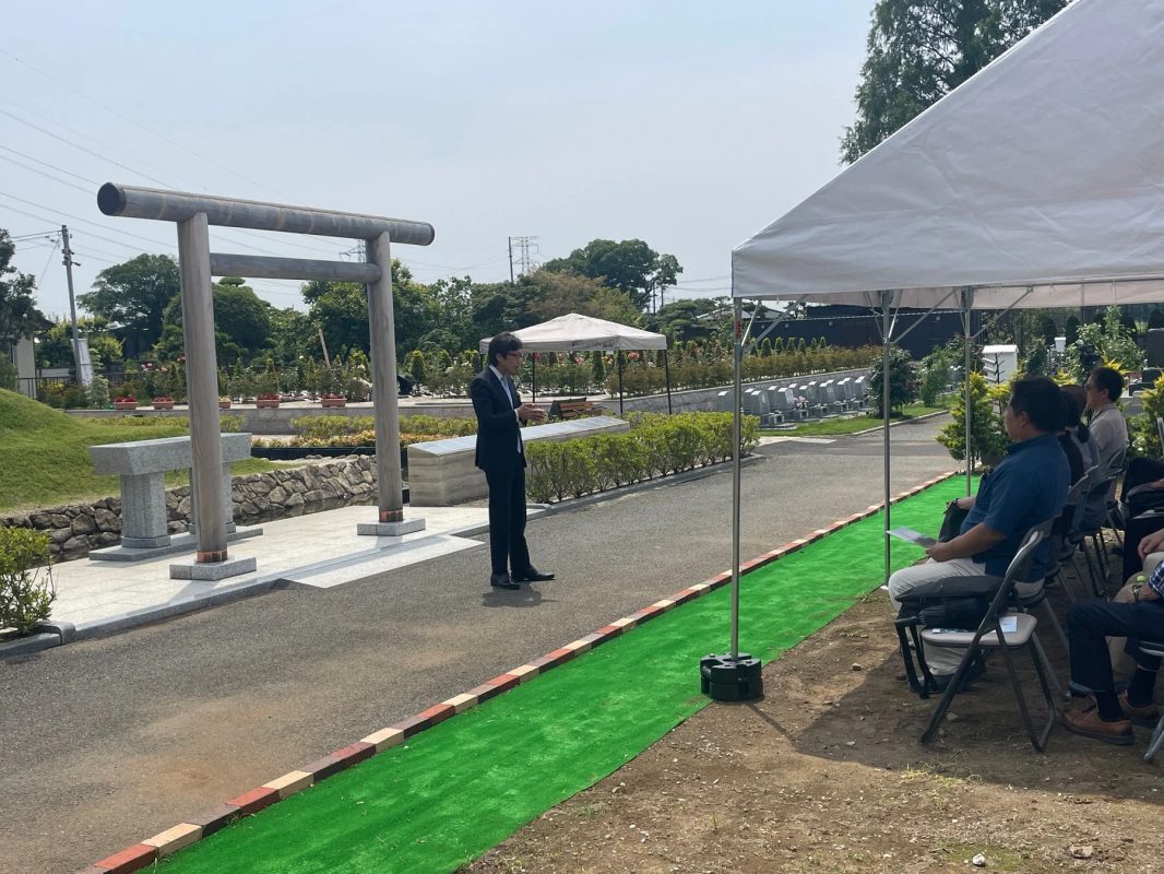 霊園または墓地のような場所で屋外イベントが開催されており、スーツ姿の男性が話している。テントの下には聴衆が座り、背景には鳥居と多くの墓石が見える。