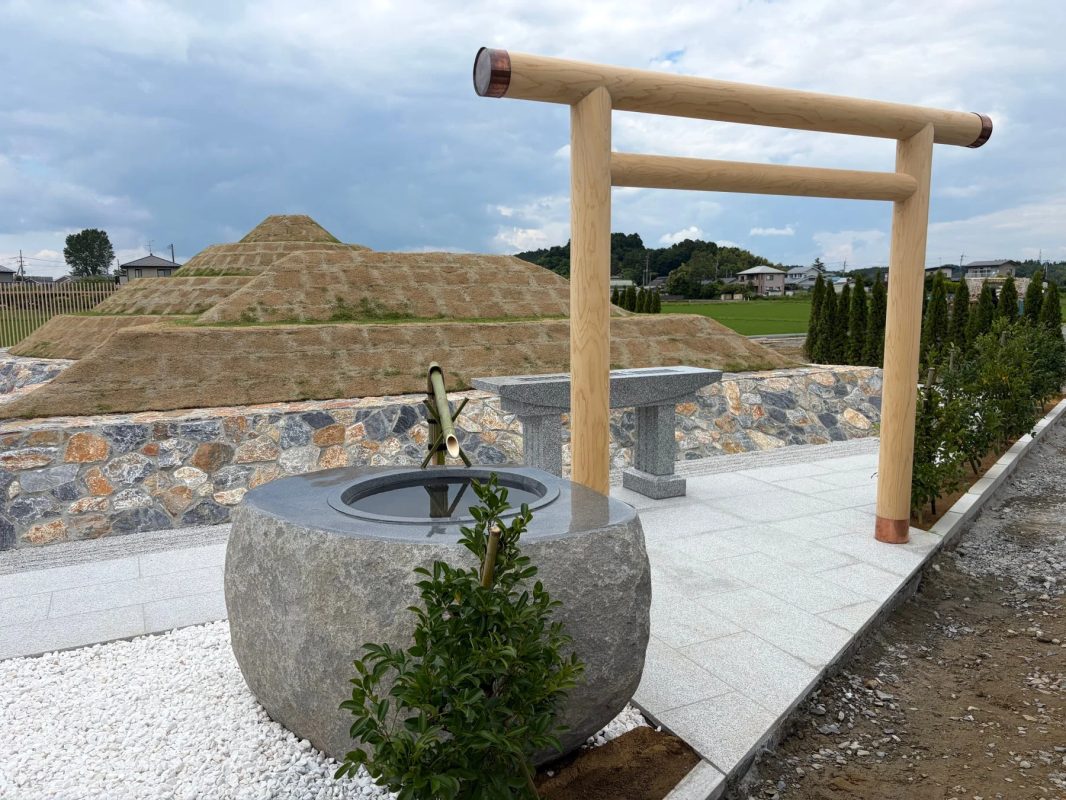 木製の鳥居と石造りの手水鉢が配置された日本の風景です。背景には段々になった芝生の大きな古墳のような盛り土が見えます。