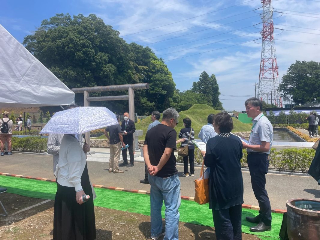 晴れた日に人々が屋外で集まり、鳥居や古墳のような丘、送電線が見える場所を訪れている。イベントか見学ツアーと思われる。