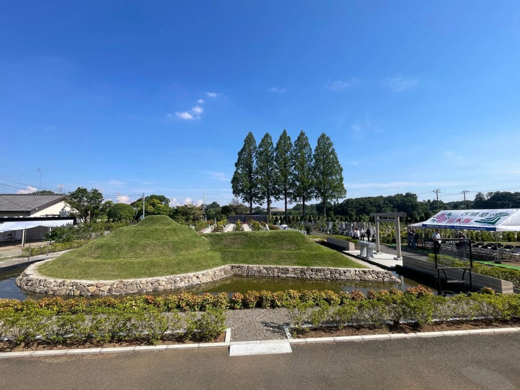 青空の下、古墳のような芝生の盛り土と水辺が特徴的な公園墓地の風景です。樹木葬や永代供養墓を提供する「野田ほたるローズガーデン」の看板が見え、現代的な供養の場であることが伺えます。