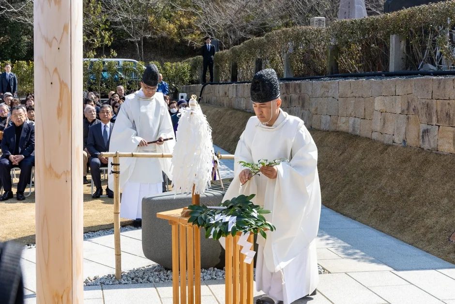 屋外で白い装束をまとった神職が、榊を捧げる神道の儀式を執り行っている様子