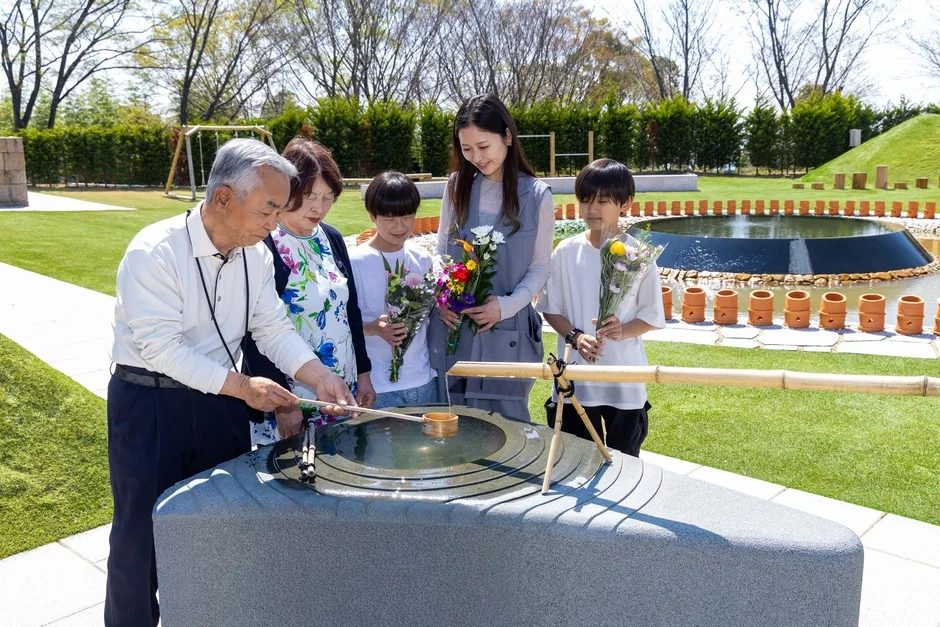 晴れた日に家族が屋外で水を使った儀式を行っている様子。高齢の男性が柄杓で水を注ぎ、他の家族が花を持って見守っている。背景には池と緑豊かな庭園が広がる。