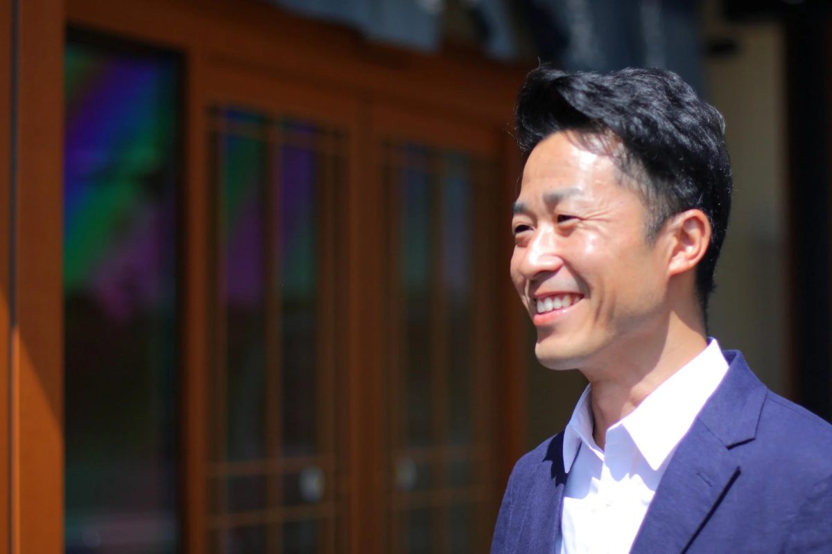 青いジャケットと白いシャツを着たアジア人男性が、明るい日差しの下で笑顔を見せているポートレートです。背景には木製の窓枠が見えます。