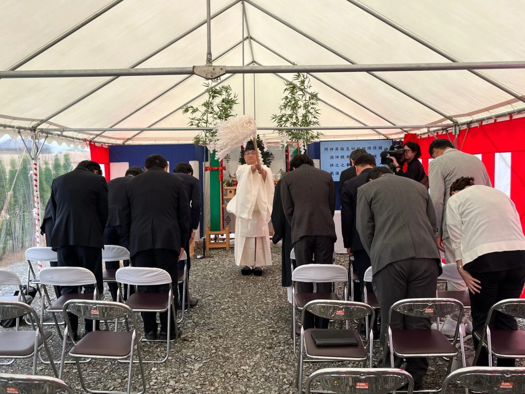 白いテントの下で神主が神事を執り行っており、スーツ姿の参列者たちが頭を下げて拝礼している様子です。地鎮祭などの伝統的な儀式が行われており、背景のスクリーンには儀式の次第が映し出されています。