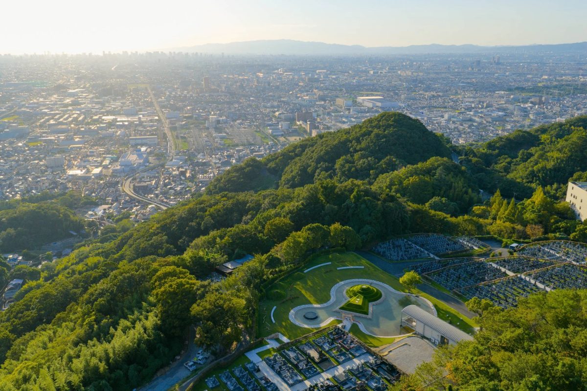高台から広がる都市のパノラマと、緑豊かな丘に位置する広大な墓苑を捉えた写真。夕暮れ時の柔らかな光が街並みと自然を美しく照らし出しています。