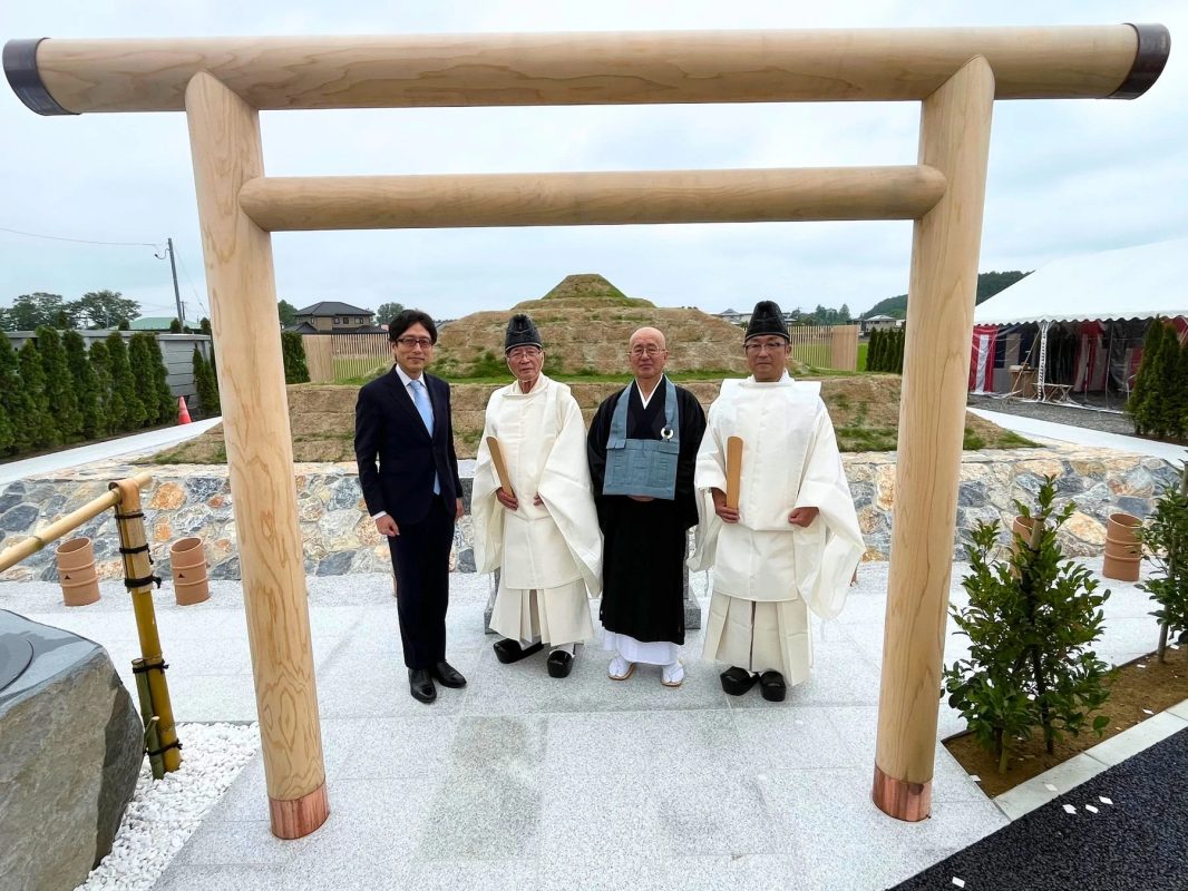 新しい木製の鳥居の下で、スーツ姿の男性と伝統的な衣装をまとった3人の男性が並んで立っている。背景には古墳のような盛り土が見える。
