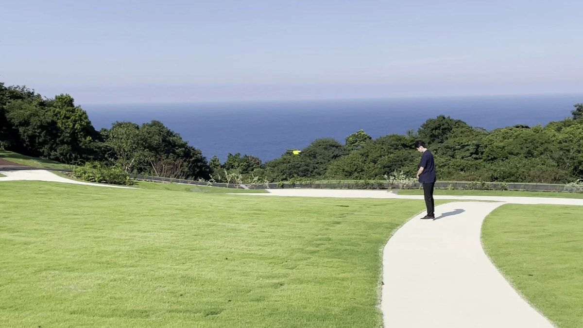 青い海と空が広がる開放的な場所で、一人の人物が芝生と小道の広がる風景の中に立っています。自然豊かな公園のような場所で、リラックスした雰囲気が感じられます。