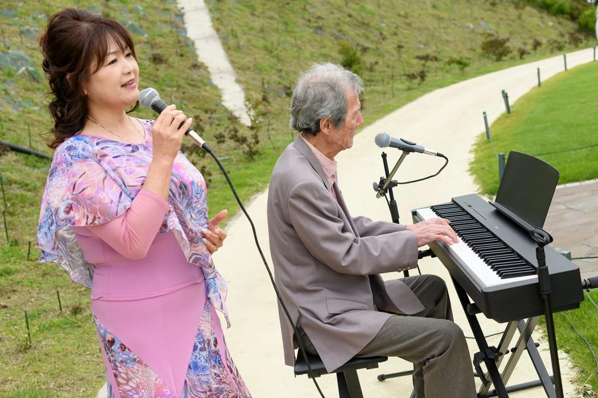 屋外で女性がマイクを持って歌い、隣で男性が電子ピアノを演奏している様子。自然豊かな場所での音楽パフォーマンスが伺えます。