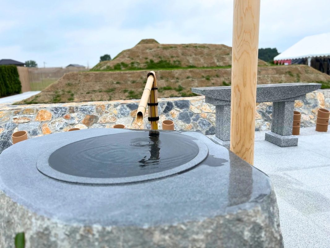 水が滴り落ちる竹筒と石の手水鉢が写っている。水面には波紋が広がり、背景には土盛りされた古墳のような景色が見える。
