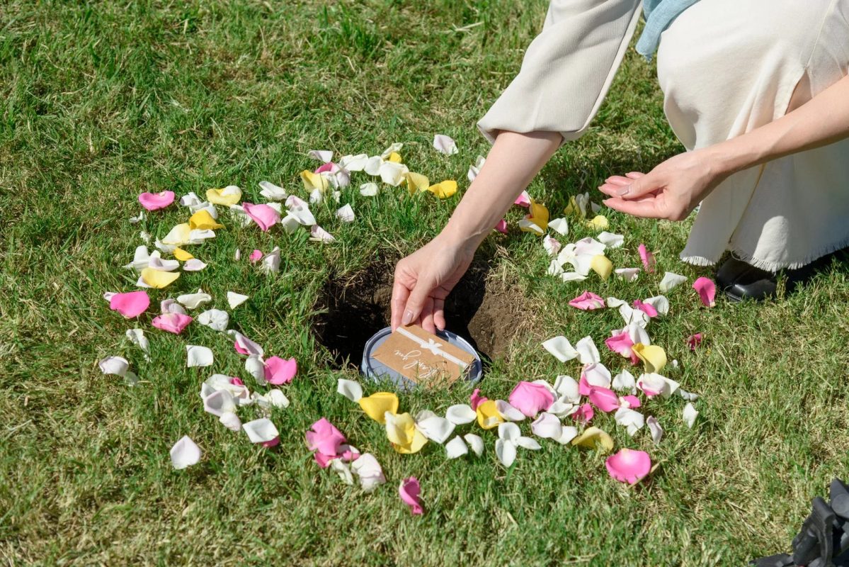 緑の芝生に開けられた小さな穴に、女性が「Thank you」と書かれたカードが乗った容器を納めている様子。周囲にはピンク、黄色、白の花びらが散らされており、自然葬や追悼の儀式を思わせる場面です。