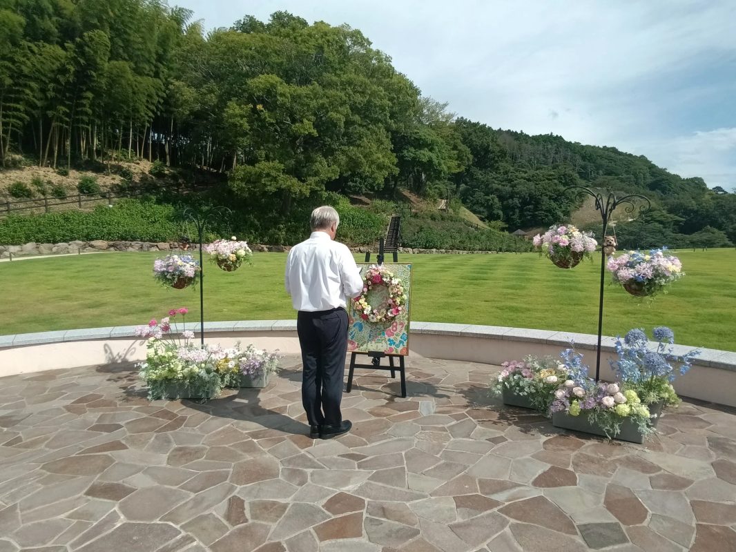 屋外の広い芝生と森を背景に、石畳の広場で一人の男性がイーゼルに立てられた花飾りの前で立っています。周囲にはたくさんの鉢植えの花が飾られており、穏やかな雰囲気のガーデンイベントの様子です。