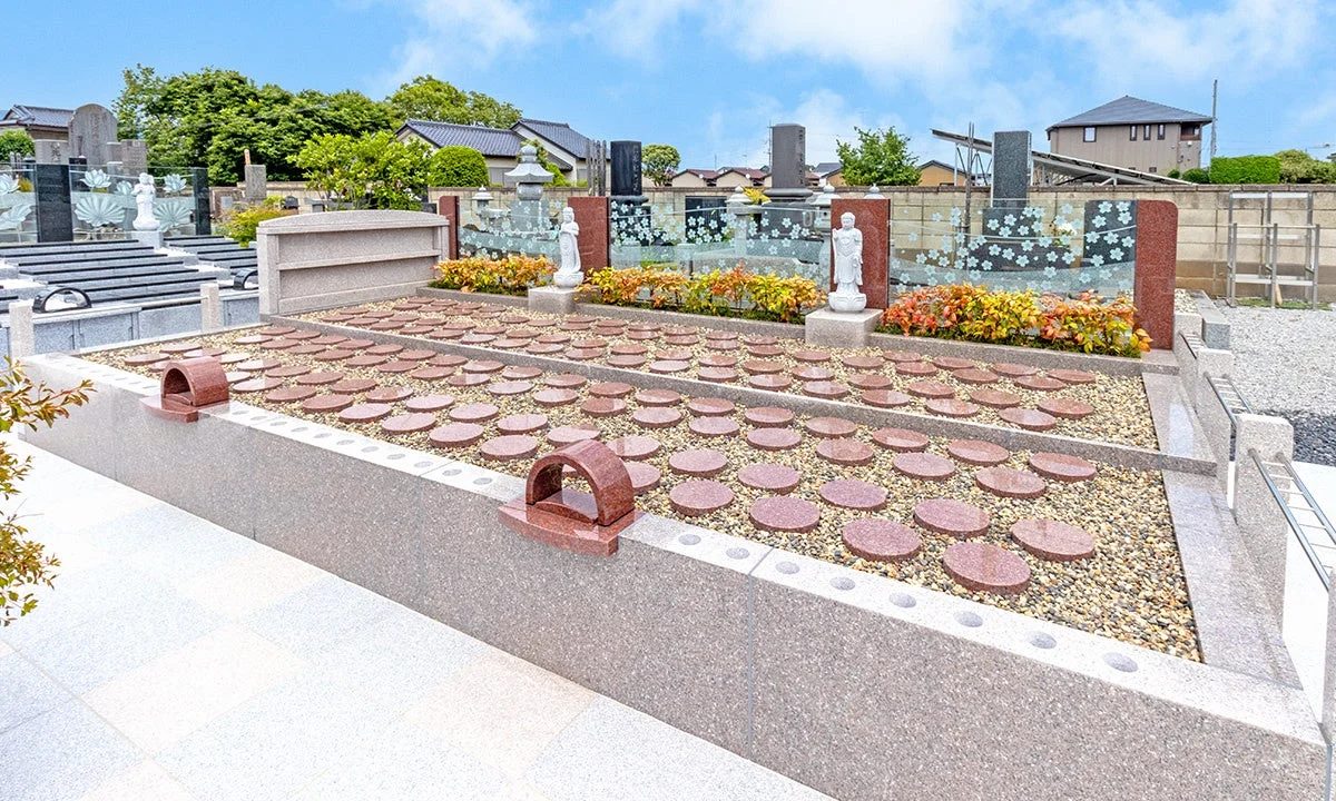 現代的な日本の墓地の風景です。砂利の上に円形の石が並べられた区画が複数あり、奥には花柄のガラスパネルや白い仏像、色とりどりの植物が配置されています。青空の下、手入れの行き届いた静かで落ち着いた空間が広がっています。