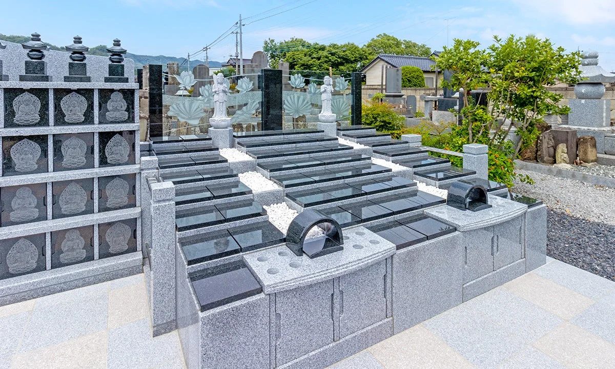 青空の下、近代的な墓地の一角が写されています。多くの花崗岩製の墓石や納骨壇が整然と並び、一部には仏像や蓮の模様が施されたガラスの仕切りも見られます。供養のための施設が並ぶ静かな風景です。