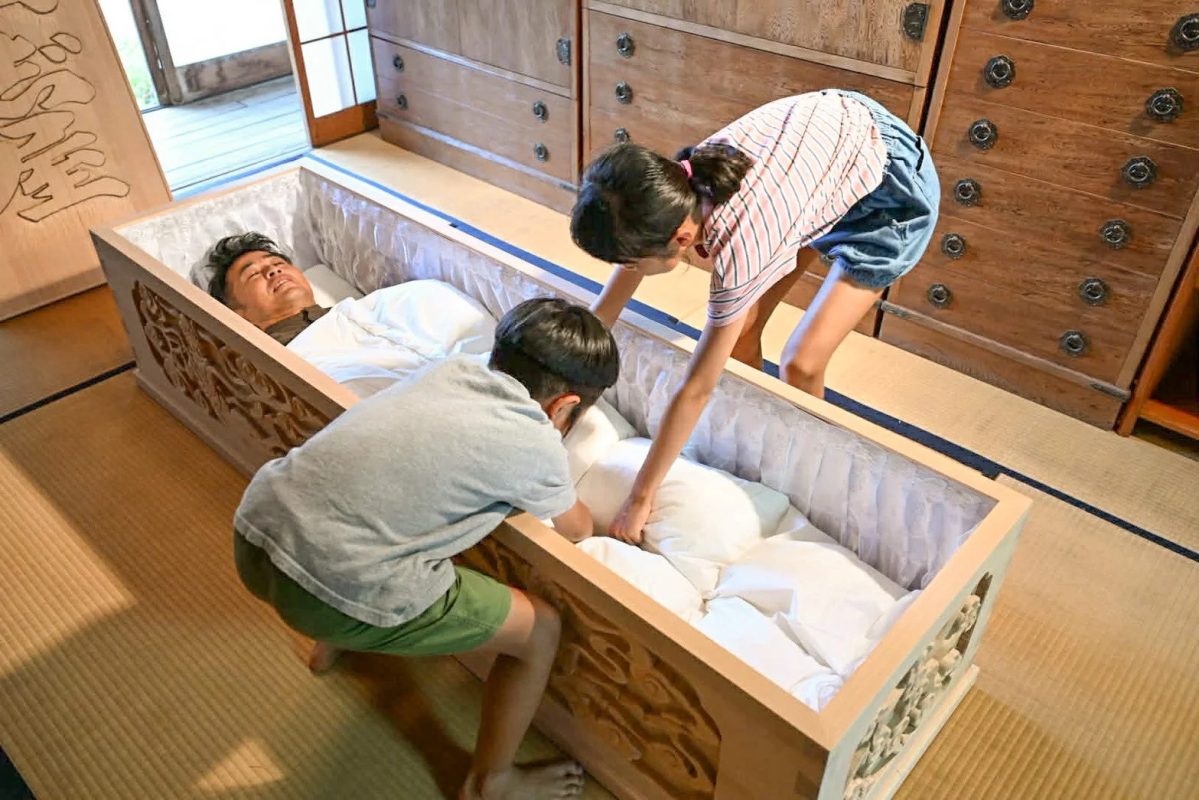 和室で男性が横たわる棺に子供が枕を整える
