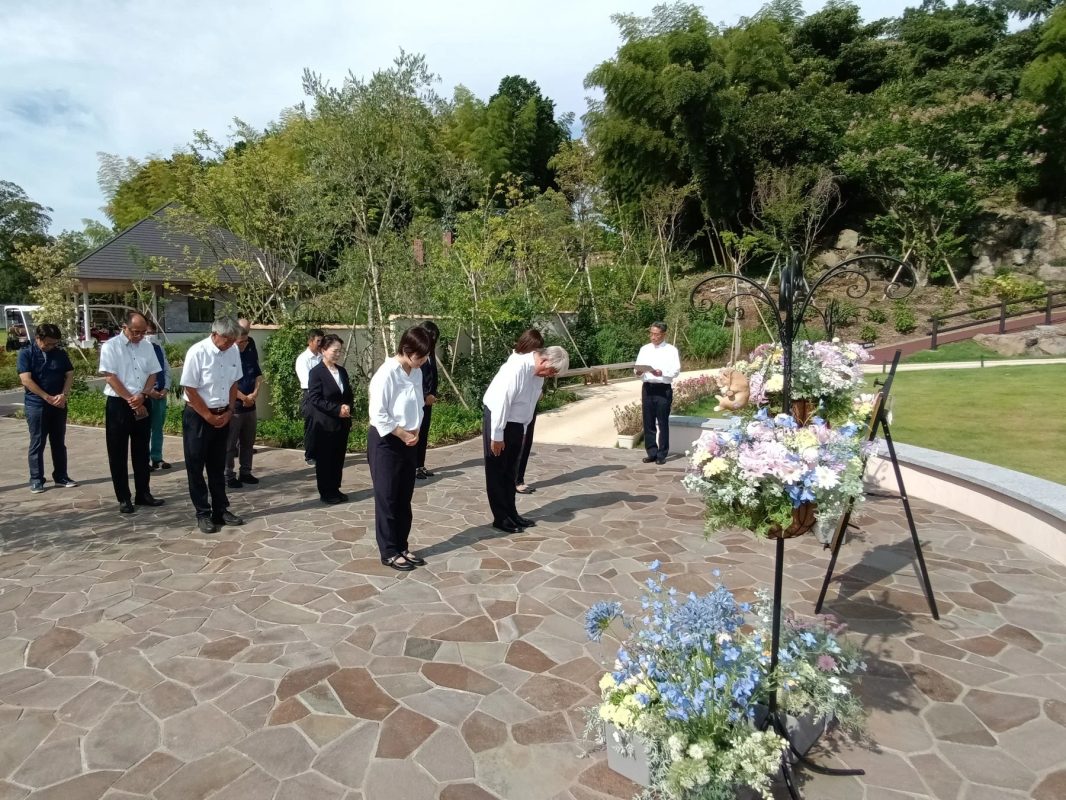 屋外の庭園で人々が集まり、一部の参加者が深くお辞儀をしている場面です。色とりどりの花が飾られており、何らかの式典や追悼行事が行われている様子がうかがえます。
