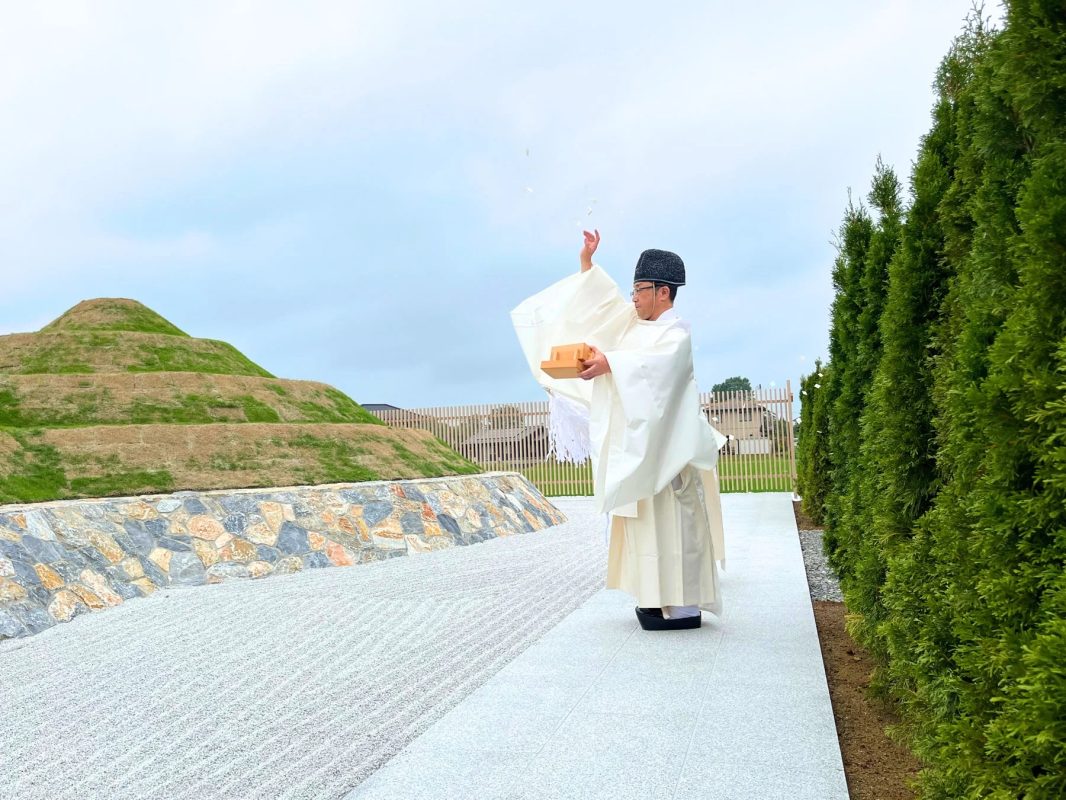 白い装束をまとった神主が、枯山水と段状の墳丘がある庭園で、上空に白い紙片を撒く儀式を行っています。