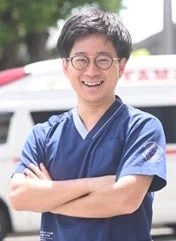 眼鏡をかけた笑顔のアジア人男性が、青いスクラブを着て腕を組み、救急車を背景に立っています。医療従事者らしきプロフェッショナルな雰囲気を醸し出しています。