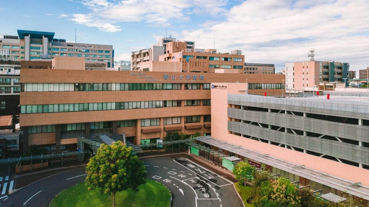 青空の下、岡山大学病院の建物と隣接する駐車場が広がる様子を捉えた航空写真です。病院の看板や駐車場の案内表示が見えます。