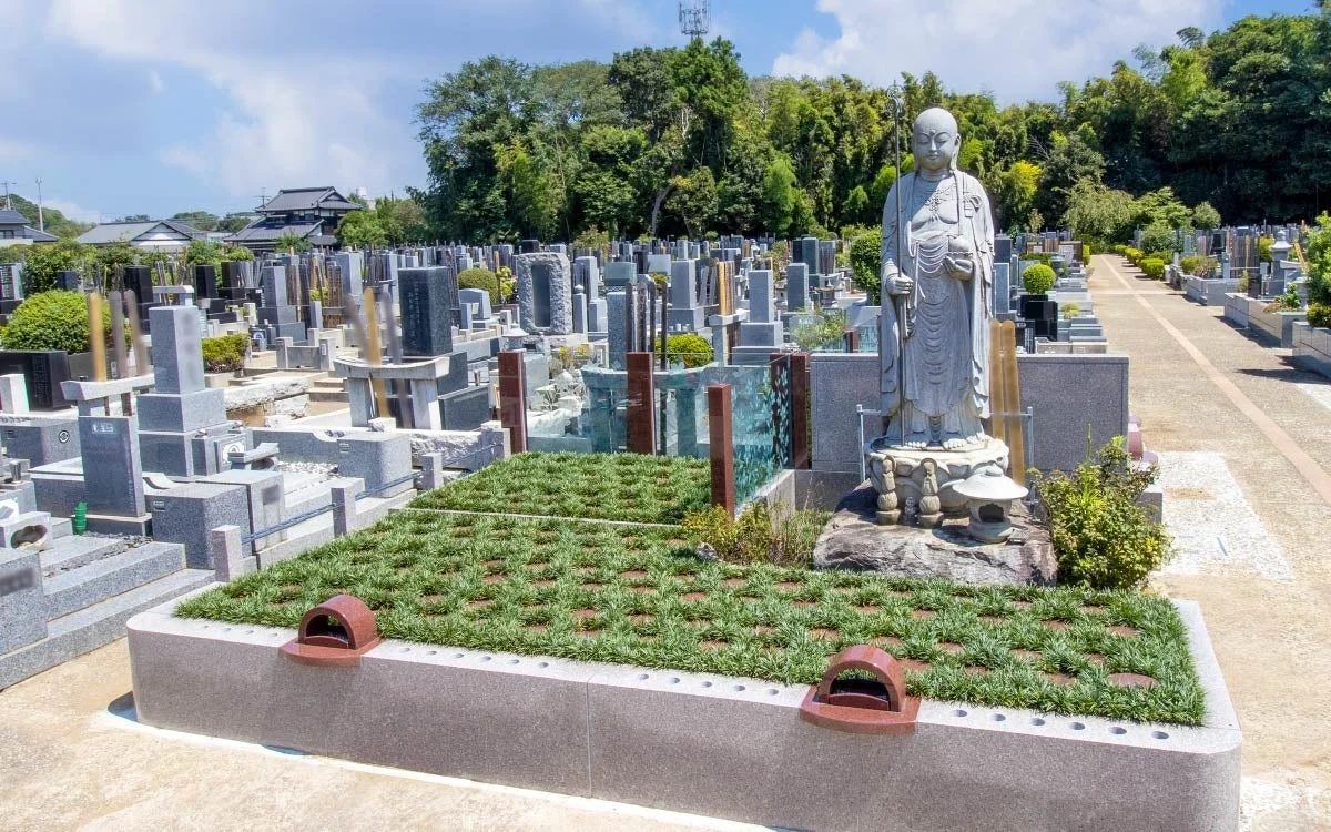 青空の下、広がる日本の墓地で、数多くの墓石が立ち並び、手前には緑の芝生が整えられた区画と、穏やかな表情の仏像が立っています。静かで厳かな雰囲気が漂っています。
