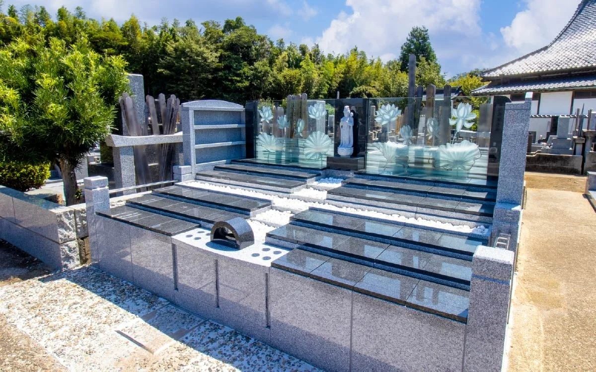 青空の下、現代的なデザインの墓地が写っています。グレーと黒の御影石で作られた墓石が並び、蓮の模様が施されたガラスパネルや仏像が特徴的です。背景には日本の寺院建築も見えます。